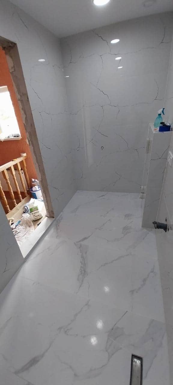 Vedere de ansamblu a băii placate cu porțelan alb aspect marmură, cu rigolă liniară în pardoseală.